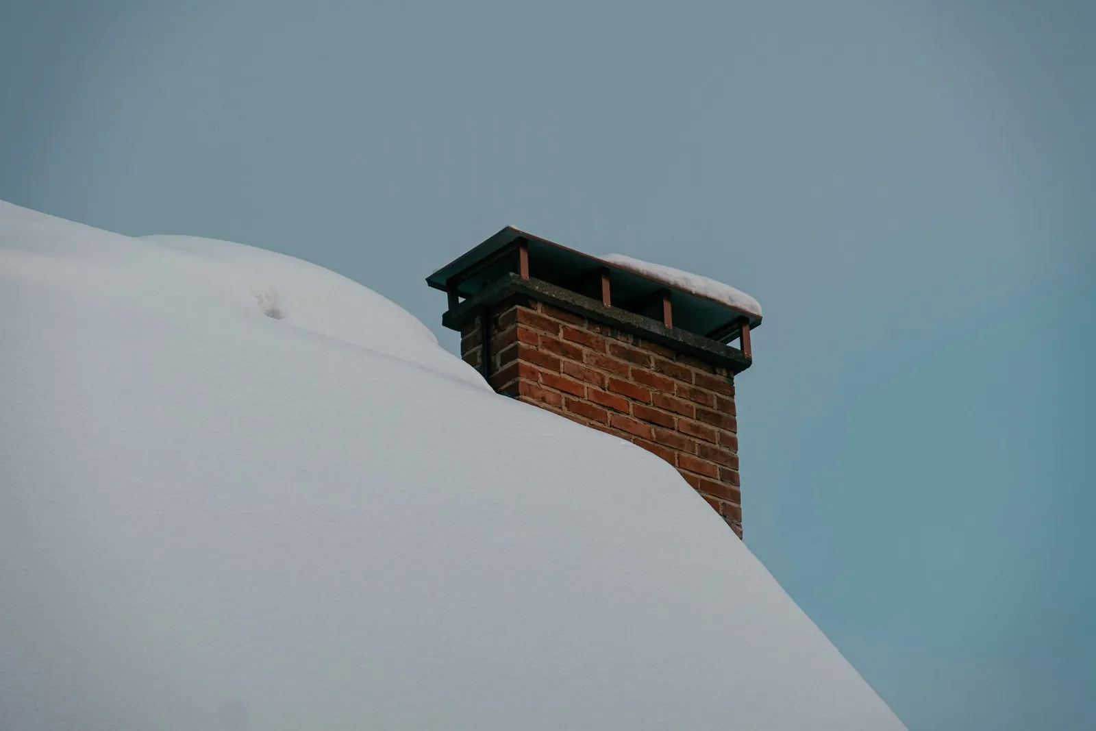 Brick chimney with a metal cap rising from a deep snow-covered roof